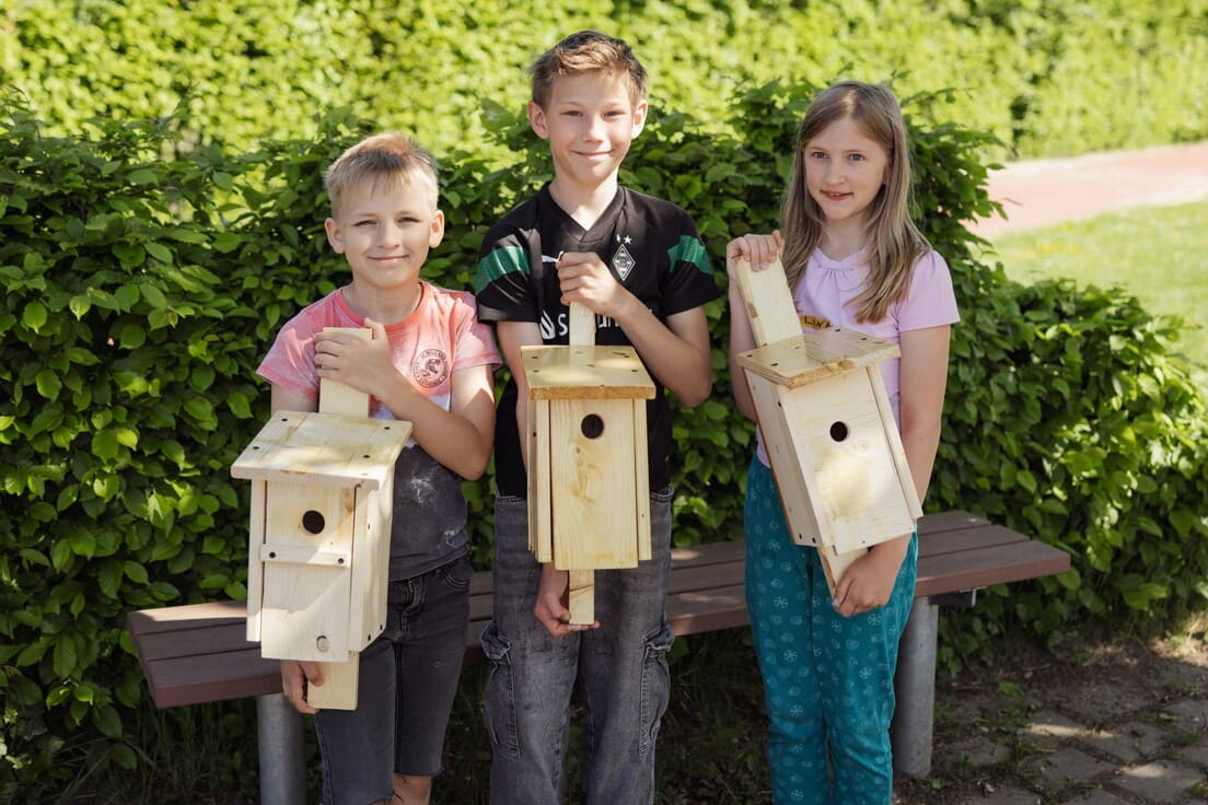 Drei Grundschulkinder stehen vor einer Bank und präsentieren die selbst gebauten Vogelhäuschen aus Holz.