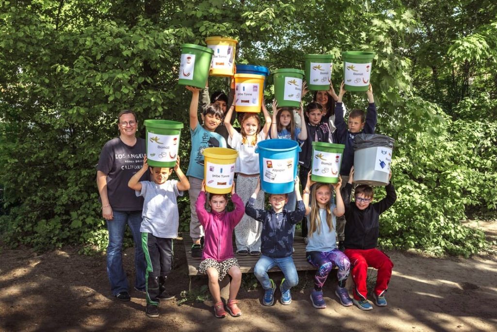 Eine Grundschulklasse sitzt bzw. steht auf einer Bank vor einer Hecke und zeigt die Mülleimer die sie zur besseren Mülltrennung an ihrer Schule gebastelt haben.
